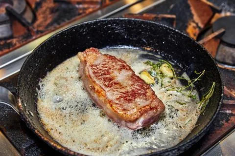 Frying pan on a stove on which a large piece of meat is fried with butter Stock Photos