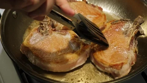Frying pork chops on a kitchen stove close-up with wooden tongs and metal pan Stock Footage 338167447