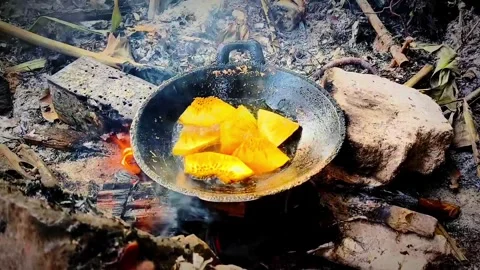 Frying sliced breadfruit pieces in a pan over a traditional wood fire Stock Footage 345181382