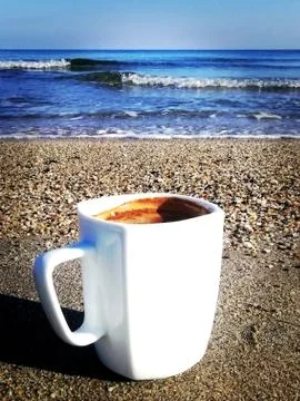 A full cup of coffee with foam stands on gray sand by the sea Stock Photos
