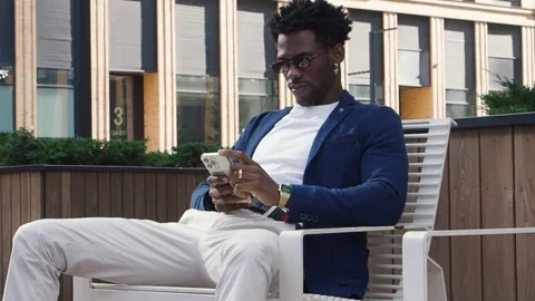 Full length black man sits on bench in urban environment and looks at phone Stock Footage 254347220