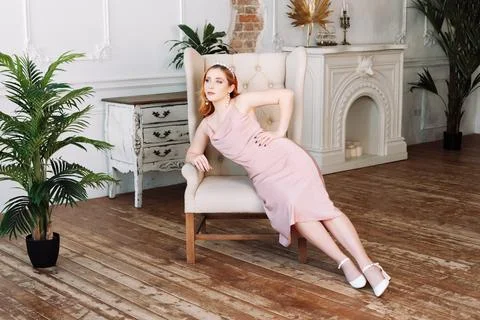 Full-length portrait of a red-haired woman in a beige silk dress sitting in a Stock Photos