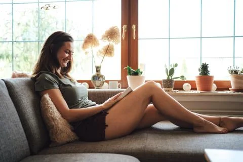 Full length side view of smiling Caucasian woman reading book on sofa near Stock Photos