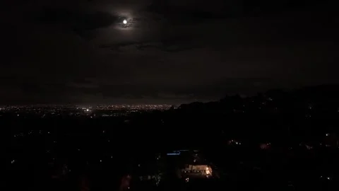 A full moon illuminating behind clouds with a lit up city below Stock Footage 310210517