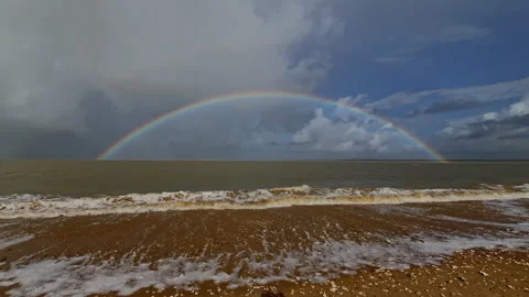 The Full rainbow stretches above a windy beach as foamy waves roll onto the Stock Footage 324806509