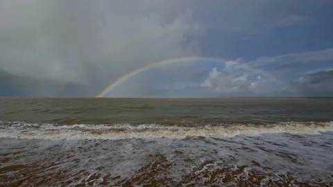 A Full rainbow stretches above a windy beach as foamy waves roll onto the sand Stock Footage 324884102