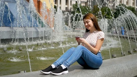 Full-size portrait of slim young girl sits near the fountain in jeans and black Video stock 99399161