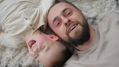 Fun daddy with beard and his little son lying at the white fur carpet and have i Stock Footage 125863738