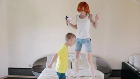 Fun Mom and son are jumping on the bed. Redhead girl singing while holding a hai Stock Footage 156300948