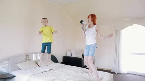 Fun Mom and son are jumping on the bed. Redhead girl singing while holding a hai Stock Footage 156300963
