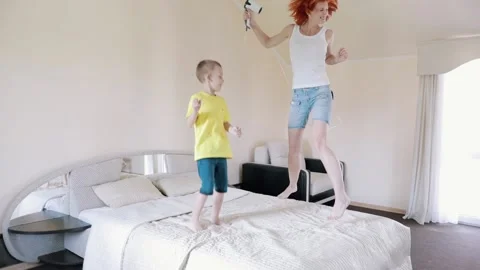 Fun Mom and son are jumping on the bed. Redhead girl singing while holding a hai Stock Footage 156300966