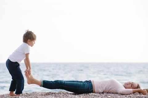 Funny active small boy lifting his mother lying on sea beach legs during spri Stock Photos