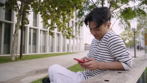 Funny Asian student guy using smartphone chatting laughing sitting on a bench - Stock Footage 221280999
