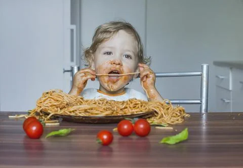 Funny baby child getting messy eating spaghetti with tomato sauce from a larg Stock Photos