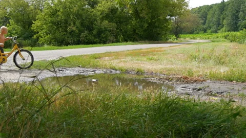 A funny barefoot five-year-old boy rides a huge puddle on a bicycle in the Stock Footage 112203178