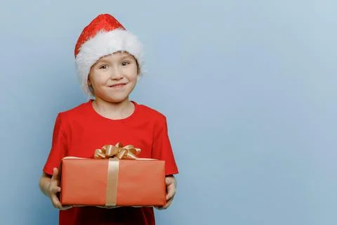Funny boy in a Santa suit and hat is holding a red gift box in his hands and Stock Photos