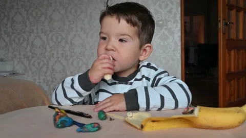 Funny caucasian toddler boy at the table eating a banana. Snack during the game Stock Footage 171381778