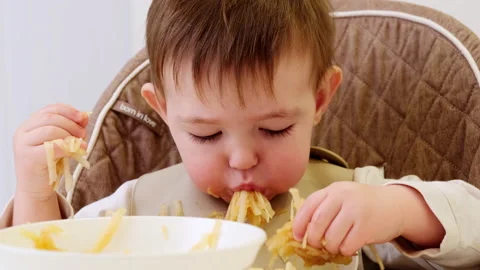 A funny child is eating a grated apple with his mouth full while sitting on a ki Stock Footage 220721950