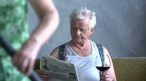 Funny elderly man drink wine and read newspaper at home on background of wife Stock Footage 63442403