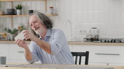 Funny enthusiastic elderly man sitting at home playing mobile game on smartphone Stock Footage 243975872