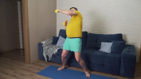 Funny fat man is actively engaged in fitness with yellow small dumbbells 스톡 동영상 169083374