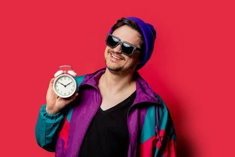 Funny guy in 80s style jacket and sunglasses holds alarm clock on red backgor Stock Photos