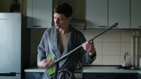 Funny guy is cleaning the kitchen. A man in a gray bathrobe plays a broom like a Stock Footage 304634334