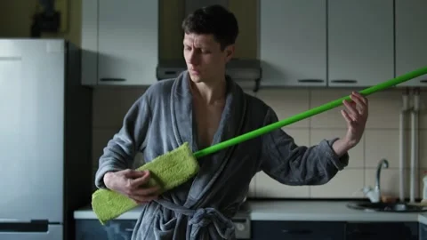 Funny guy is cleaning the room. A man in a gray bathrobe plays a mop like a rock Stock Footage 304632972