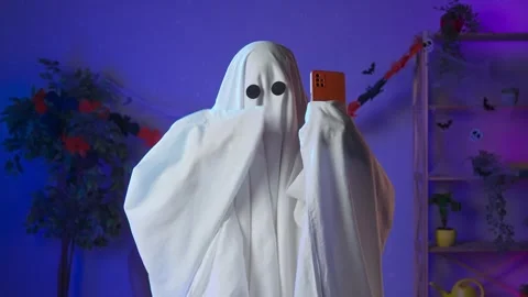 Funny halloween ghost in white sheet with black eyes in dark room taking selfie Stock Footage 285742398