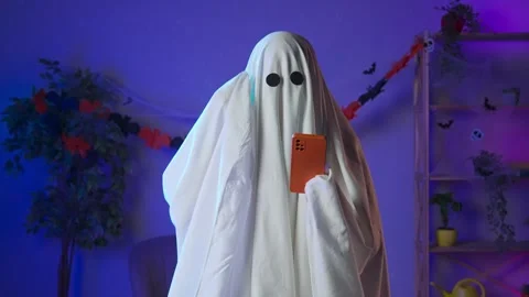 Funny halloween ghost in white sheet with black eyes in dark room texting on Stock Footage 285742478