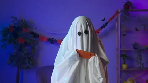 Funny halloween ghost in white sheet with black eyes in dark room playing video Stock Footage 285742651