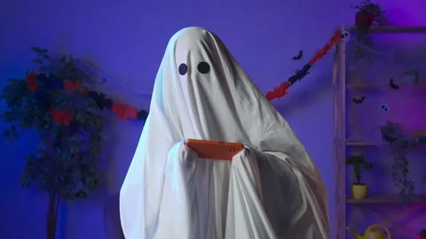 Funny halloween ghost in white sheet with black eyes in dark room playing video Stock Footage 285742681