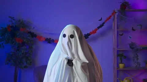 Funny halloween ghost in white sheet with black eyes in dark room plays video Stock Footage 285744020