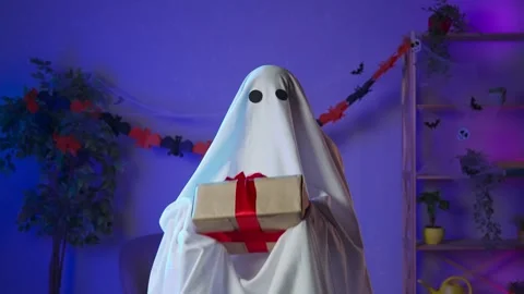 Funny halloween ghost in white sheet with black eyes in dark room holding gift Stock Footage 285744045
