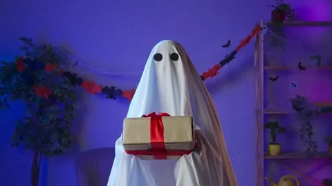 Funny halloween ghost in white sheet with black eyes in dark room holding gift Stock Footage 285744100