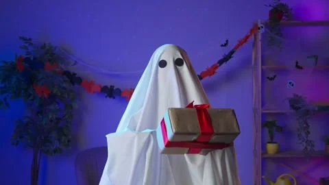 Funny halloween ghost in white sheet with black eyes in dark room holding gift Stock Footage 285744111