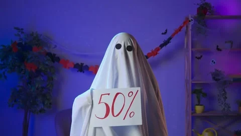 Funny halloween ghost in white sheet with black eyes in dark room shows paper Stock Footage 285744534