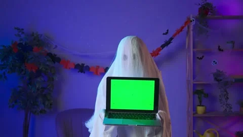Funny halloween ghost in white sheet with black eyes in dark room holding laptop Stock Footage 285746138
