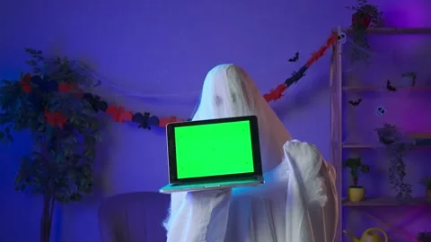 Funny halloween ghost in white sheet with black eyes in dark room holding laptop Stock Footage 285746156