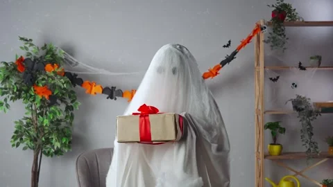 Funny halloween ghost in white sheet with black eyes in bright room holding gift Stock Footage 285746803