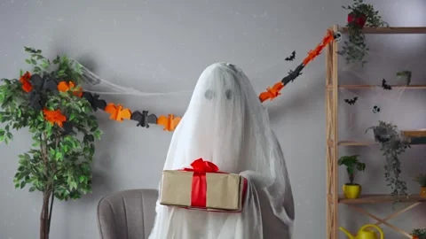 Funny halloween ghost in white sheet with black eyes in bright room holding gift Stock Footage 285746811