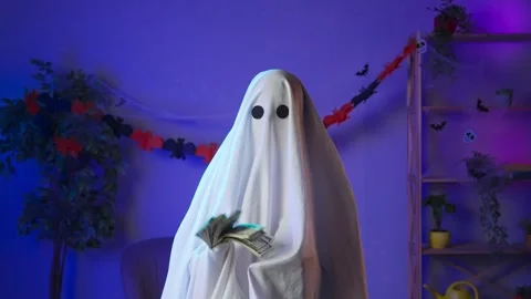 Funny halloween ghost in white sheet in dark decorated room holding fan of money Stock-Footage 285754211