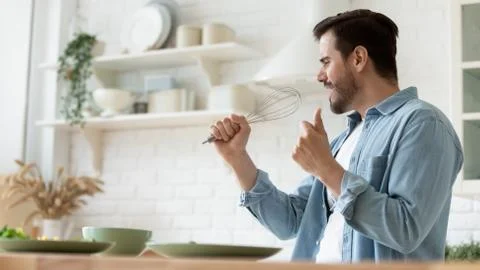 Funny handsome man singing into whist, having fun in kitchen Stock Photos