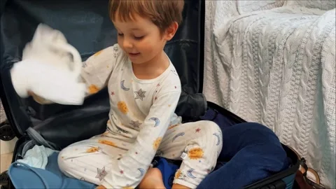 Funny kid in pajamas got into a packed suitcase and throws mom's clothes. Boy Stock Footage 255796953