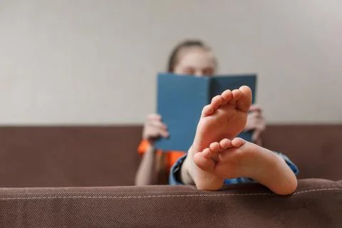 A funny little caucasian girl with bare feet with book while sitting on a sof Stock Photos