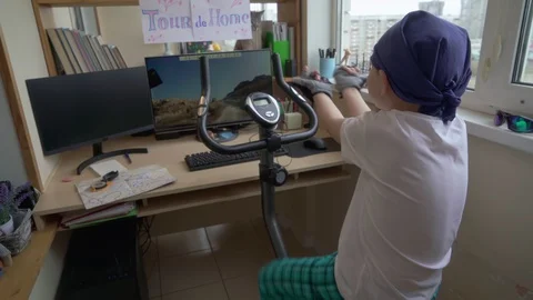 Funny Little Cyclist. Boy Is Practicing At Home On Exercise Bike And Playing Video stock 129586603