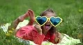 Funny little girl in big sunglasses in the shape of hearts lies on green grass HD Footage