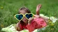 Funny little girl in big sunglasses in the shape of hearts lies on green grass HD Footage