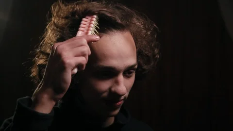 Funny man combing his long curly hair with hairbrush in the dark room Vidéo 99050517