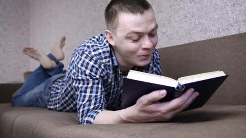 Funny man reading book while lying on couch at home. Knowledge and learning Stock Footage 129663341